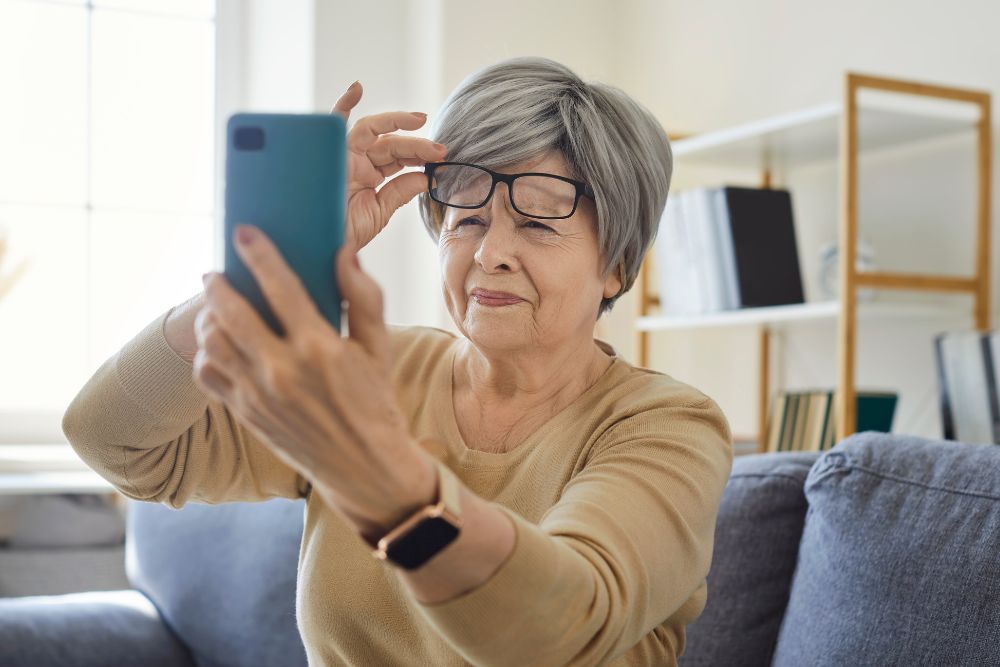 Older woman holding glasses while looking at a smartphone, representing vision changes from macular degeneration.