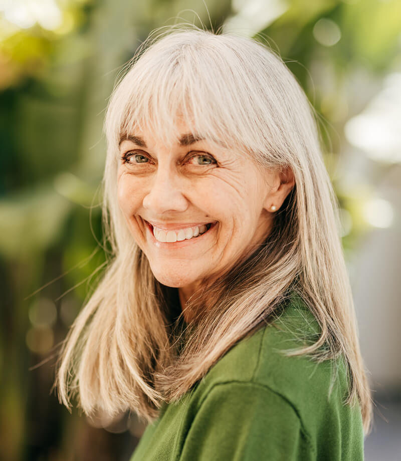 Smiling older woman outdoors with long gray hair wearing a green top.